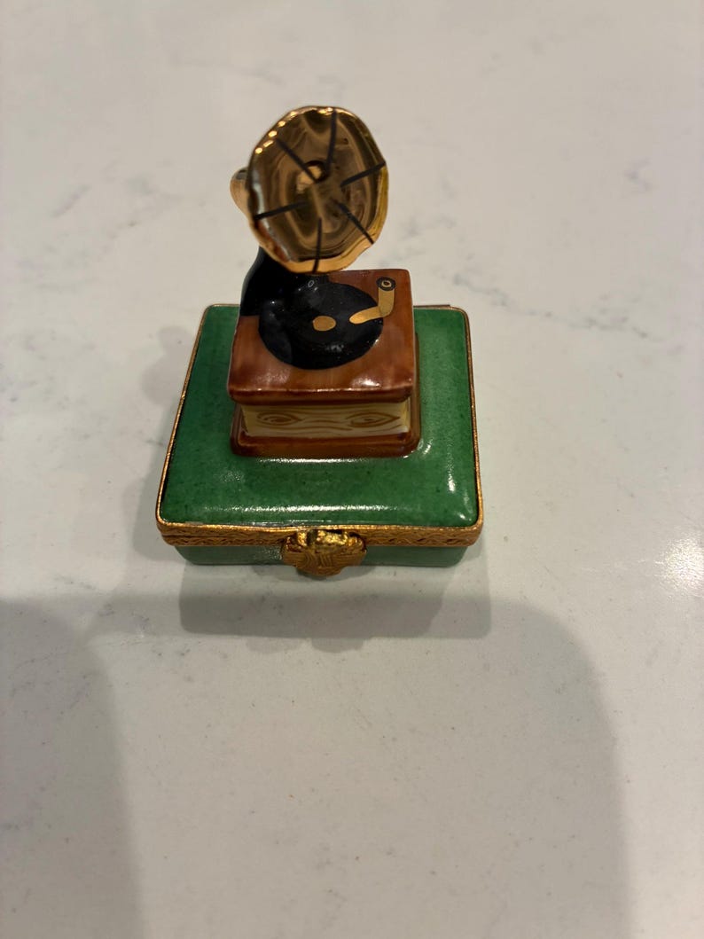 May include: A small decorative box with a green, rectangular base and gold-coloured trim. A detailed miniature gramophone sits on top, with a black record and a gold horn. The box is likely ceramic or porcelain.