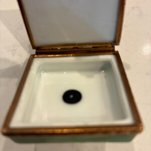 May include: A small, open trinket box with a white interior and a black dot in the centre. The box has a gold-coloured trim and a pale green exterior. The lid is open, showing the white interior.
