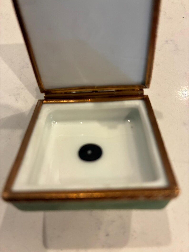 May include: A small, open trinket box with a white interior and a black dot in the centre. The box has a gold-coloured trim and a pale green exterior. The lid is open, showing the white interior.