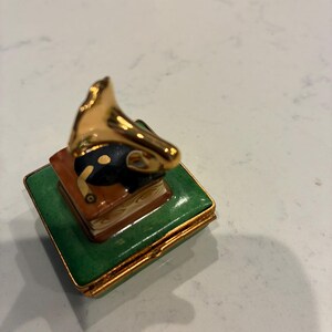 May include: A small decorative box with a gold-coloured gramophone on top of a stack of books. The box is green with gold trim. The gramophone has a black horn and gold accents. The box is likely ceramic or porcelain.