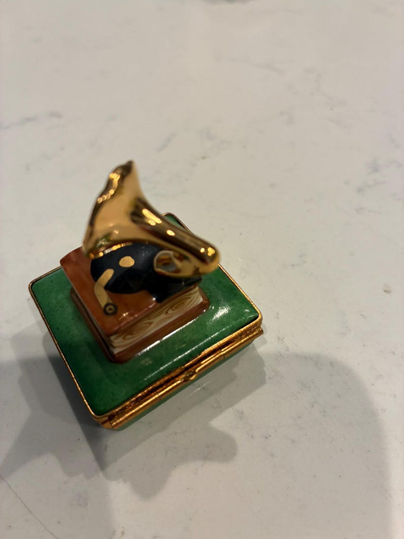 May include: A small decorative box with a gold-coloured gramophone on top of a stack of books. The box is green with gold trim. The gramophone has a black horn and gold accents. The box is likely ceramic or porcelain.