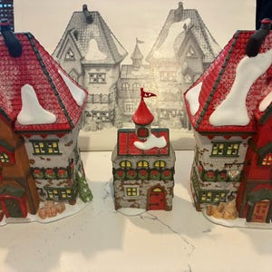 May include: Three ceramic Christmas village buildings featuring red shingled roofs with white snow accents. The left building is labeled 'North Pole Dolls', the center is a small tower, and the right building is labeled 'Santa's Bear Works'.