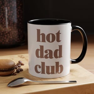 May include: White ceramic mug with a black interior and handle. The mug has the text "hot dad club" in brown. A spoon and a macaron are on a wooden surface beside the mug.