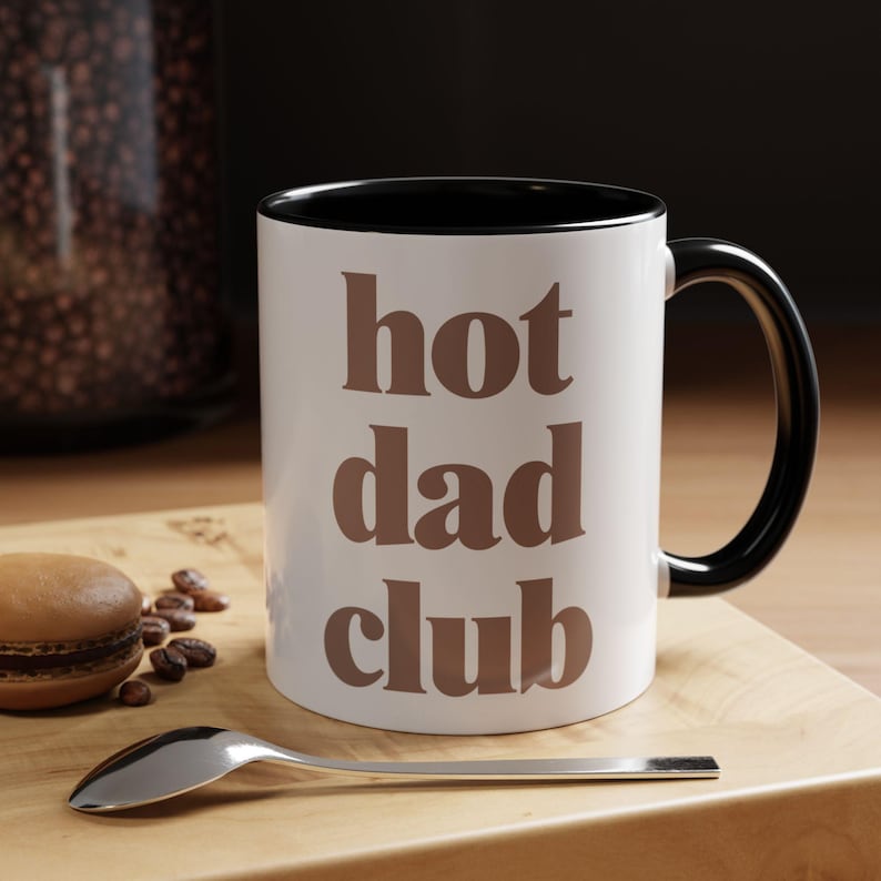 May include: White ceramic mug with a black interior and handle. The mug has the text "hot dad club" in brown. A spoon and a macaron are on a wooden surface beside the mug.