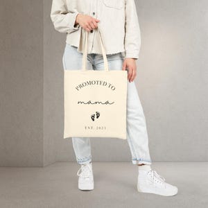 May include: Cream-colored tote bag with the words "PROMOTED TO mama" in black script. Below the text are two black footprints and the text "EST. 2025". The bag is held by a person wearing light-wash jeans and white sneakers.