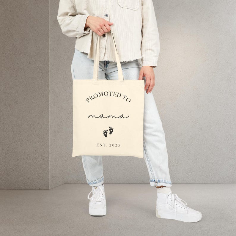 May include: Cream-colored tote bag with the words "PROMOTED TO mama" in black script. Below the text are two black footprints and the text "EST. 2025". The bag is held by a person wearing light-wash jeans and white sneakers.