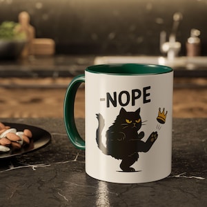 May include: White ceramic mug with a green handle and interior. The mug features a black cat illustration with a crown and the word "-NOPE" in black. The mug is sitting on a black countertop.