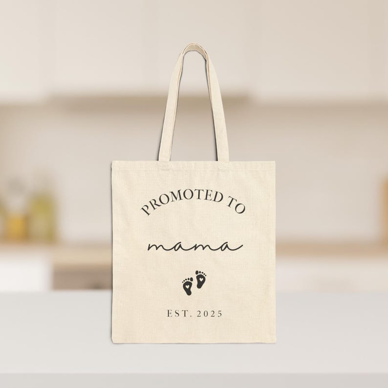 May include: Cream-colored tote bag with the text "PROMOTED TO mama" in a mix of fonts, along with two small footprints and the year "EST. 2025" printed in black. The bag has matching cream-colored handles.