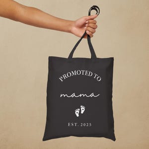 May include: Black tote bag with the text "PROMOTED TO mama" in white script, along with two white baby footprints and the year "EST. 2025". The bag is held by a hand.