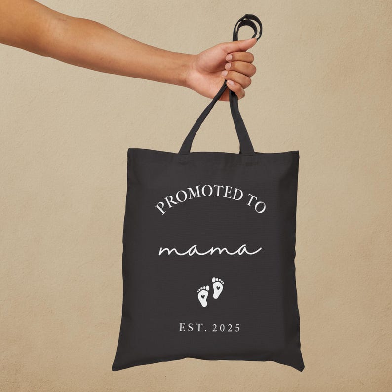 May include: Black tote bag with the text "PROMOTED TO mama" in white script, along with two white baby footprints and the year "EST. 2025". The bag is held by a hand.