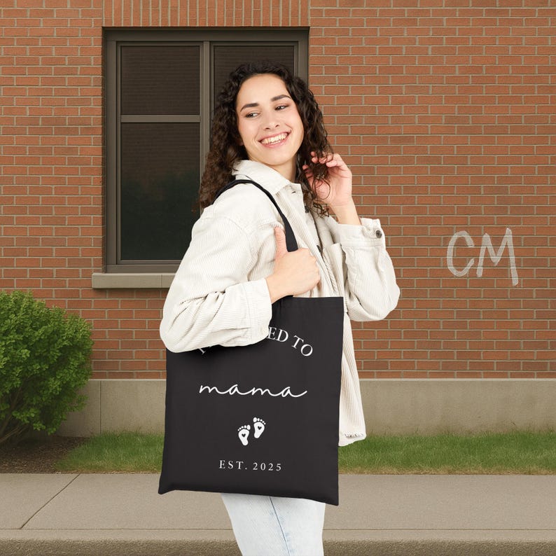 May include: Black tote bag with white text that reads "I'm headed to mama" in a script font, with baby footprints and "EST. 2025" below. The bag is carried by a person wearing a white jacket.