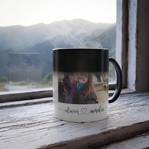 May include: A black and white color-changing mug with a photograph of a couple kissing. The mug has the names "oliver" and "amelia" with a heart symbol. The mug is sitting on a white wooden surface.