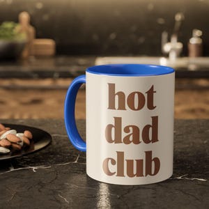 May include: A white ceramic mug with a blue interior and handle. The mug has the words "hot dad club" in brown lettering. The mug is on a black countertop.