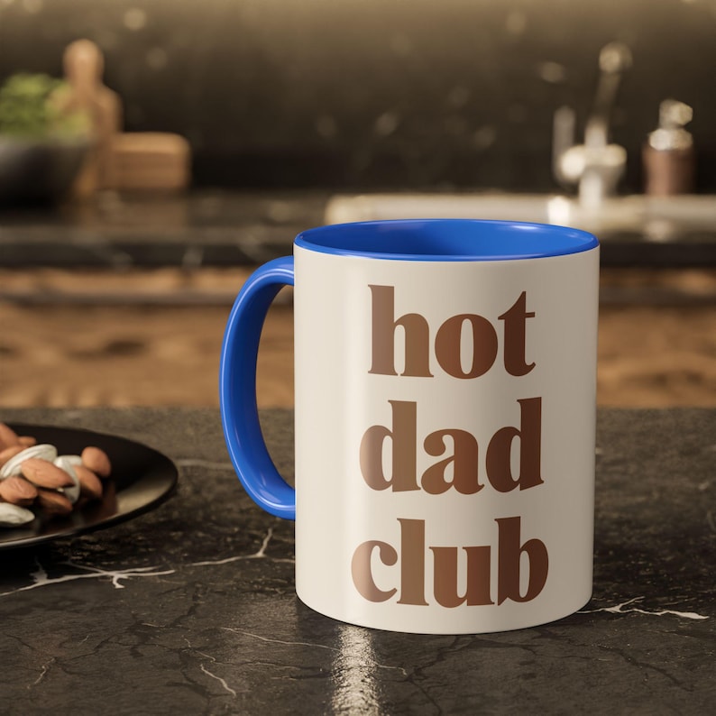 May include: A white ceramic mug with a blue interior and handle. The mug has the words "hot dad club" in brown lettering. The mug is on a black countertop.