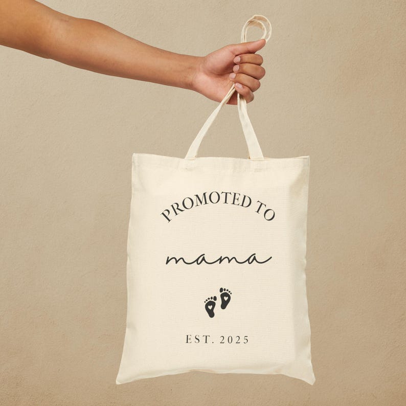 May include: Cream-colored tote bag with the words "PROMOTED TO mama" in black script. Below the text are two black baby footprints and the year "EST. 2025". The bag is held by a person.