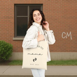 May include: Cream-colored tote bag with the words "I'm going to be a mama" in black script, along with two black footprint graphics and the text "EST. 2025". The bag is held by a person.