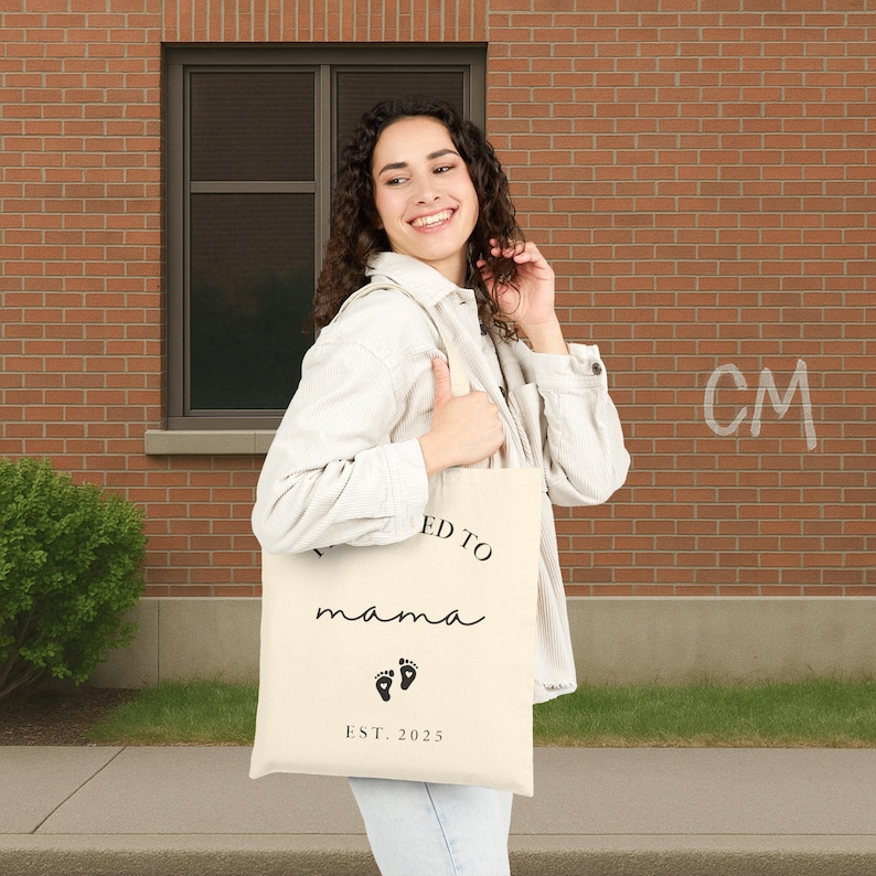May include: Cream-colored tote bag with the words "I'm going to be a mama" in black script, along with two black footprint graphics and the text "EST. 2025". The bag is held by a person.