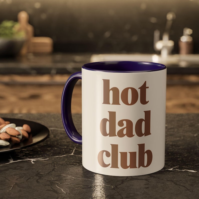 May include: A white ceramic mug with a dark blue interior and handle. The mug features the words "hot dad club" in brown block letters. The mug is on a black countertop.