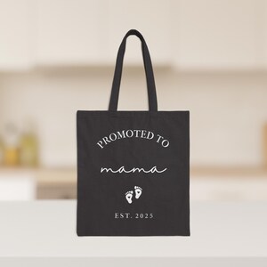 May include: Black tote bag with white text that reads "PROMOTED TO mama" in a script font, with two baby footprints and "EST. 2025" below. The bag has two handles and is on a white surface.