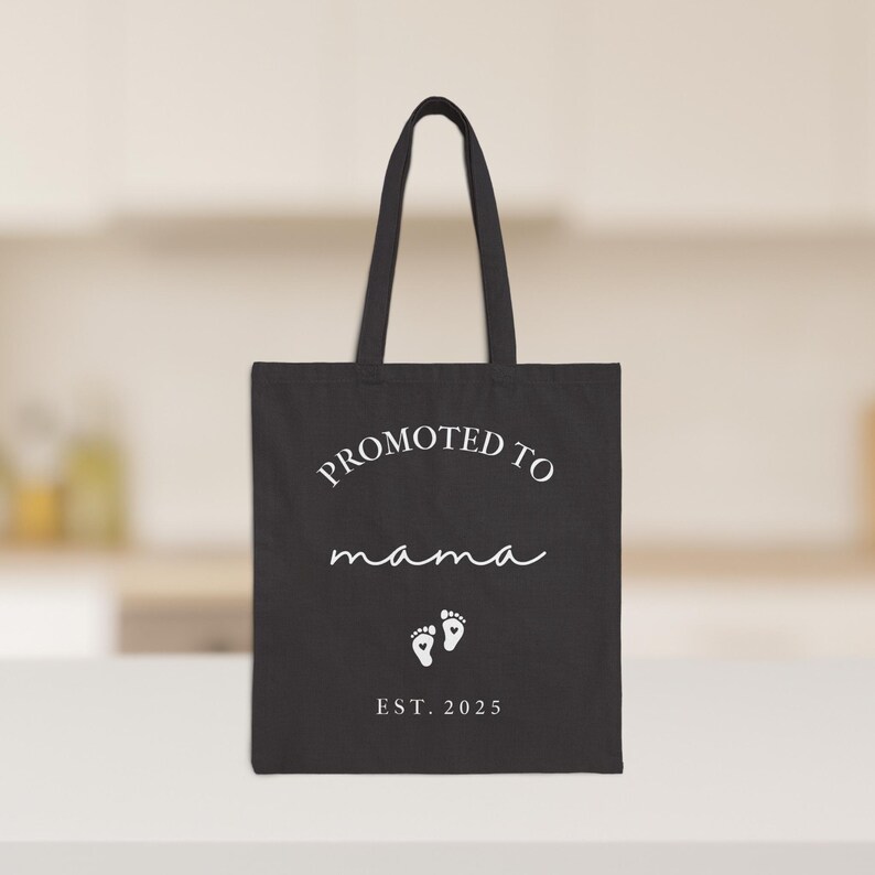 May include: Black tote bag with white text that reads "PROMOTED TO mama" in a script font, with two baby footprints and "EST. 2025" below. The bag has two handles and is on a white surface.