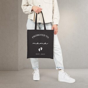 May include: Black tote bag with the text "PROMOTED TO mama" in white script. Below the text are two white baby footprints and the text "EST. 2025". The bag has black handles.