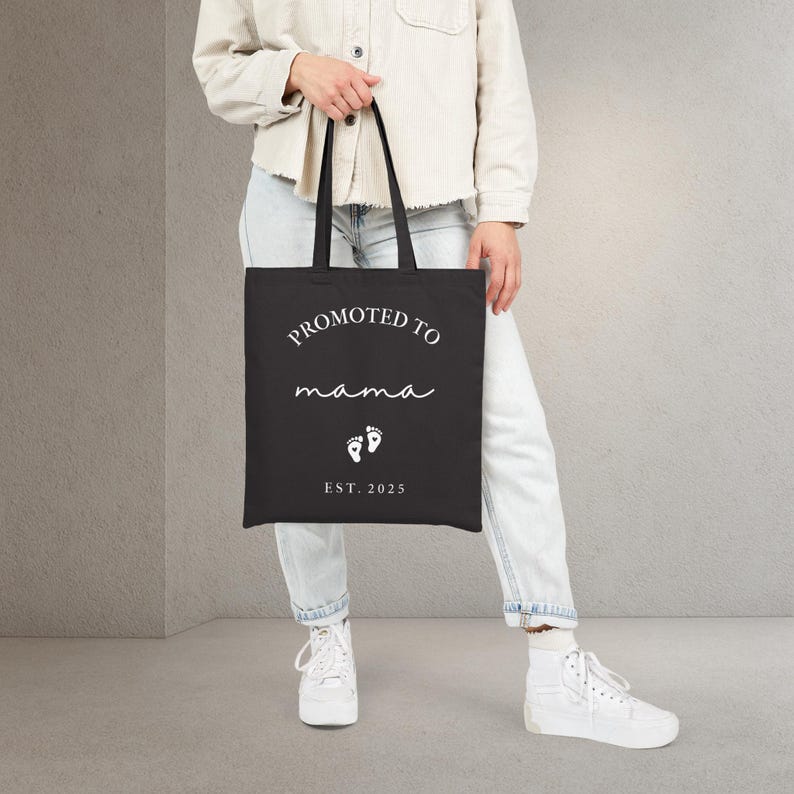May include: Black tote bag with the text "PROMOTED TO mama" in white script. Below the text are two white baby footprints and the text "EST. 2025". The bag has black handles.