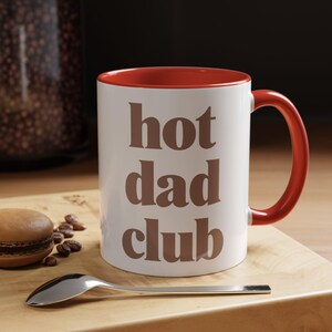 May include: White ceramic mug with a red interior and handle. The mug has the text "hot dad club" in brown letters. The mug is on a wooden board with a spoon, a macaron, and coffee beans.