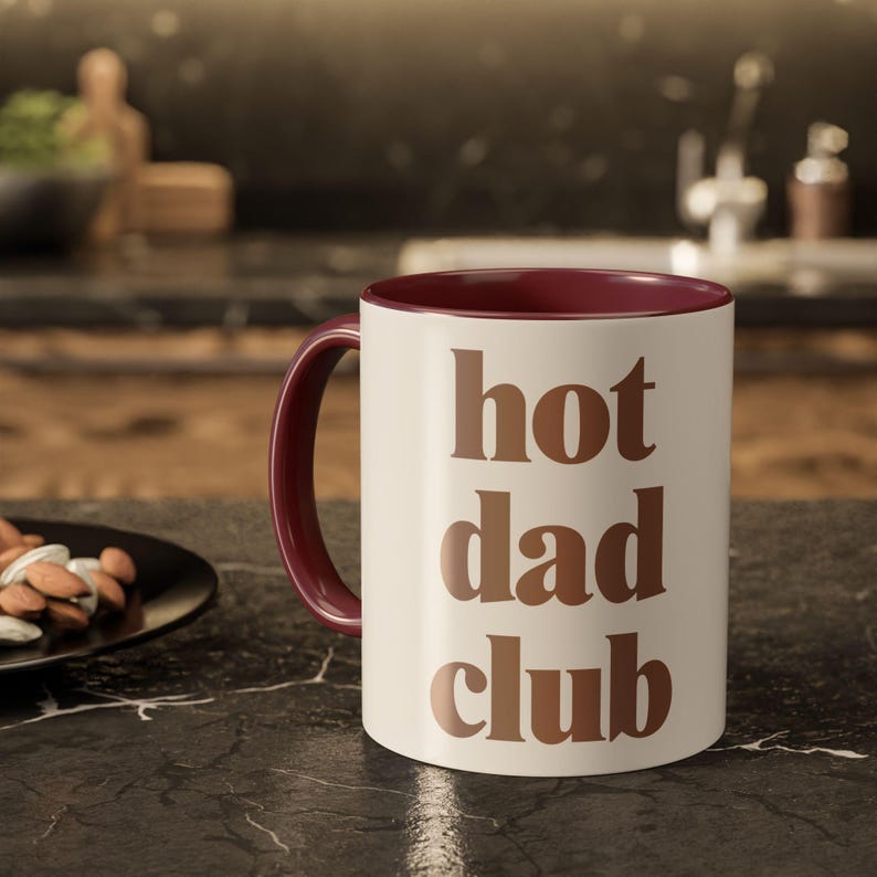 May include: Cream-coloured ceramic mug with a burgundy interior and handle. The mug features the words "hot dad club" in brown block letters. The mug is on a dark countertop.