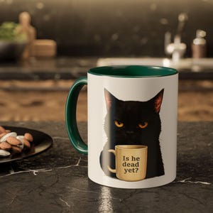 May include: White ceramic mug with a green interior and handle, featuring a black cat illustration with yellow eyes. The mug within the illustration has the text "Is he dead yet?" printed on it. The mug is on a dark surface.