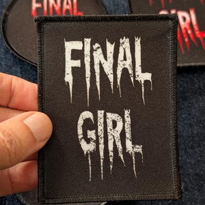 May include: Black square patch with the words "FINAL GIRL" in white, dripping font. Other patches with the word "FINAL" in red are also visible. The patches are on a blue denim background.