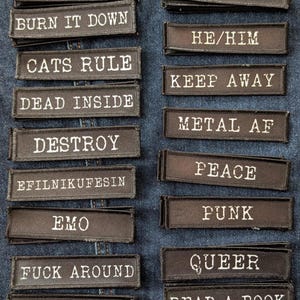 May include: A collection of black rectangular patches with white embroidered text. Phrases include "Breathe," "Burn It Down," "Cats Rule," "Dead Inside," "Destroy," "Emo," "Fuck Around," and "Find Out." Other patches say "Final Girl," "He/Him," "Keep Away," "Metal AF," "Peace," "Punk," "Queer," and "Read a Book."