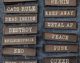 Puede incluir: Un conjunto de parches rectangulares negros con texto bordado en blanco. Las frases incluyen "Breathe", "Burn It Down", "Cats Rule", "Dead Inside", "Destroy", "Emo", "Fuck Around" y "Find Out". Otros parches dicen "Final Girl", "He/Him", "Keep Away", "Metal AF", "Peace", "Punk", "Queer" y "Read a Book."