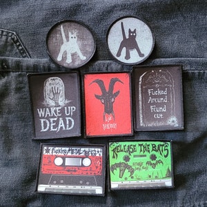 May include: A collection of eight iron-on patches featuring various designs, including a cat, a goat, a tombstone, and a cassette tape. The patches are black, white, red, and green.