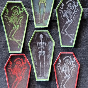 May include: A collection of six coffin-shaped patches. Each patch features a skeleton design in various colors, including green, red, and white, with a black background. The patches have a green or red border.