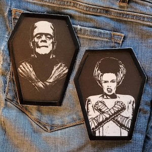 May include: Two black and white iron-on patches in the shape of coffins. The patch on the left features a black and white image of Frankenstein's monster with his arms crossed. The patch on the right features a black and white image of the Bride of Frankenstein with her arms crossed.