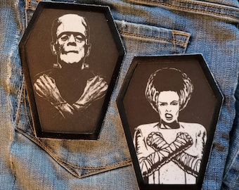 May include: Two black and white iron-on patches in the shape of coffins. The patch on the left features a black and white image of Frankenstein's monster with his arms crossed. The patch on the right features a black and white image of the Bride of Frankenstein with her arms crossed.
