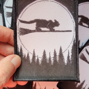 May include: Square patch with a black squirrel riding a broom in front of a full moon and a forest silhouette. The patch has a black border and is likely an iron-on or sew-on patch for clothing or accessories.