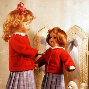 May include: Two dolls wearing red jackets and plaid skirts stand next to each other. The doll on the left has curly blonde hair and a red bow. The doll on the right has straight red hair and a red bow. Both dolls are holding a small white dog.