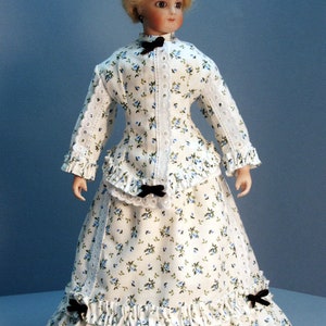 K&ouml;nnte beinhalten: Eine Porzellanpuppe, die ein wei&szlig;es Kleid mit Blumenmuster und schwarzen Schleifenakzenten tr&auml;gt. Das Kleid hat einen ger&uuml;schten Rock und einen Spitzenbesatz.