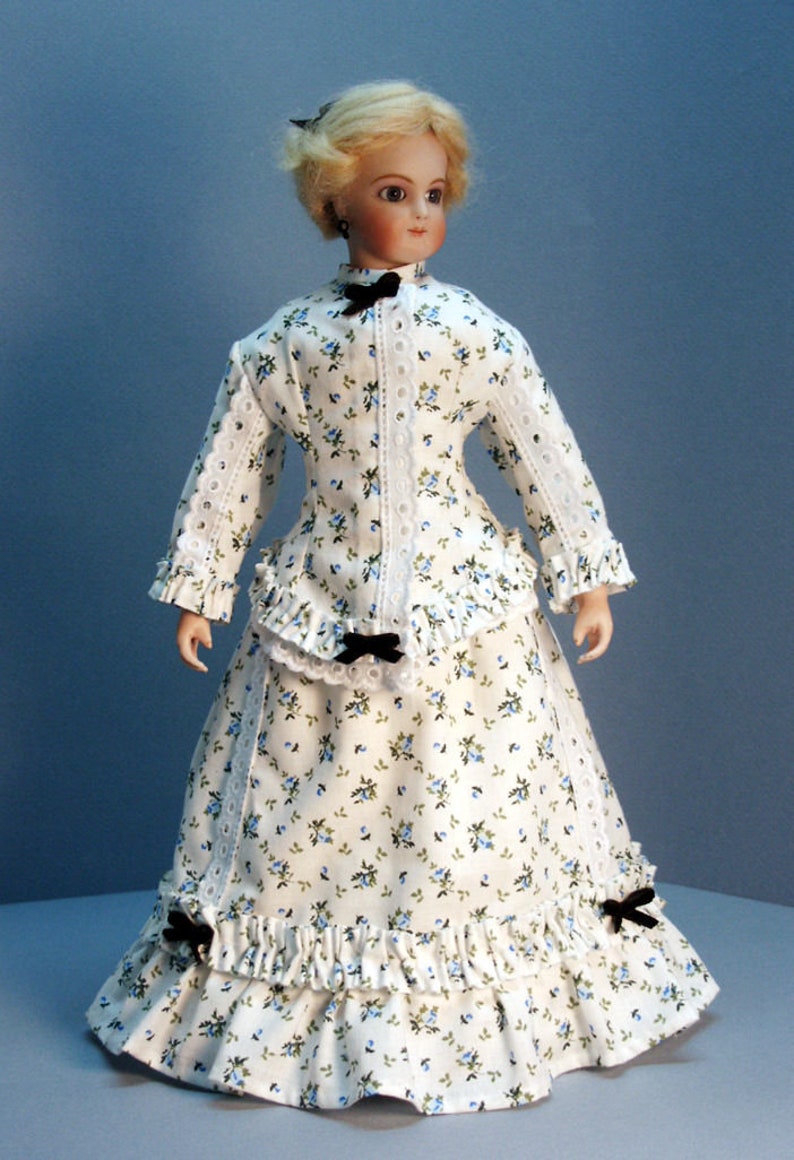 K&ouml;nnte beinhalten: Eine Porzellanpuppe, die ein wei&szlig;es Kleid mit Blumenmuster und schwarzen Schleifenakzenten tr&auml;gt. Das Kleid hat einen ger&uuml;schten Rock und einen Spitzenbesatz.