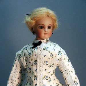 K&ouml;nnte beinhalten: Eine Porzellanpuppe mit blonden Haaren, die ein wei&szlig;es Kleid mit blauem Blumendruck und wei&szlig;en Spitzenbesatz tr&auml;gt. Die Puppe hat schwarze Augen und tr&auml;gt schwarze Ohrringe.