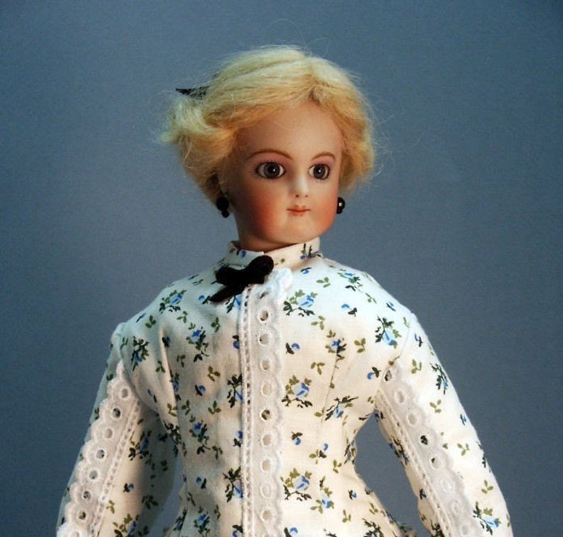 K&ouml;nnte beinhalten: Eine Porzellanpuppe mit blonden Haaren, die ein wei&szlig;es Kleid mit blauem Blumendruck und wei&szlig;en Spitzenbesatz tr&auml;gt. Die Puppe hat schwarze Augen und tr&auml;gt schwarze Ohrringe.