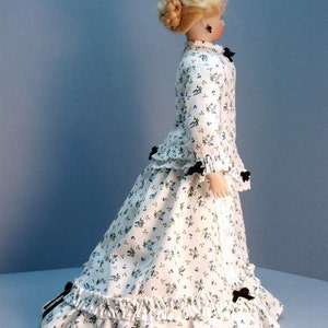 K&ouml;nnte beinhalten: Eine Porzellanpuppe, die ein wei&szlig;es Kleid mit Blumenmuster und schwarzen Schleifen tr&auml;gt. Das Kleid hat einen ger&uuml;schten Rock und ein tailliertes Oberteil. Die Puppe hat blondes Haar, das zu einem Knoten mit einer schwarzen Schleife gesteckt ist.
