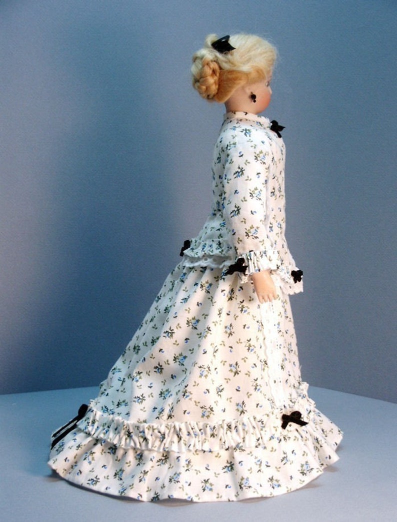 K&ouml;nnte beinhalten: Eine Porzellanpuppe, die ein wei&szlig;es Kleid mit Blumenmuster und schwarzen Schleifen tr&auml;gt. Das Kleid hat einen ger&uuml;schten Rock und ein tailliertes Oberteil. Die Puppe hat blondes Haar, das zu einem Knoten mit einer schwarzen Schleife gesteckt ist.