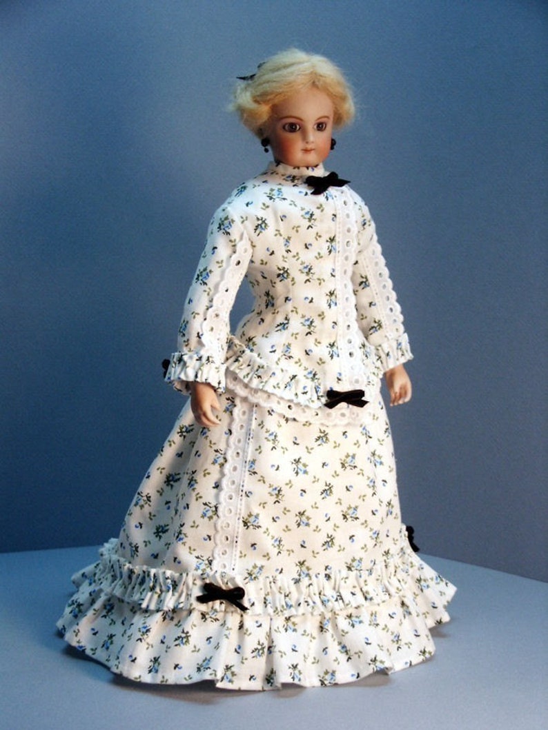 K&ouml;nnte beinhalten: Eine Porzellanpuppe, die ein wei&szlig;es Kleid mit blauem Blumenmuster tr&auml;gt. Das Kleid hat einen ger&uuml;schten Rock und eine schwarze Schleife an der Taille. Die Puppe hat blonde Haare und blaue Augen.