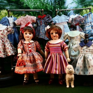 May include: Two dolls wearing colorful dresses stand in front of a rack of doll clothes. The dolls have long brown hair and are wearing red and white striped dresses. The doll clothes are a variety of colors and patterns, including floral, striped, and polka dot.
