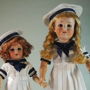 May include: Two porcelain dolls dressed in sailor outfits. The doll on the left is wearing a white sailor dress with blue trim and a white sailor hat with the word "Atlantic" on it. The doll on the right is wearing a white sailor dress with blue trim and a white sailor hat with a blue band.