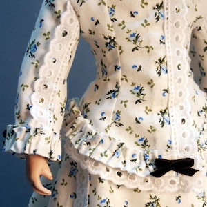K&ouml;nnte beinhalten: Ein wei&szlig;es Kleid mit blauem Blumenmuster und wei&szlig;em Spitzenbesatz. Das Kleid hat ein tailliertes Oberteil und einen vollen Rock. Das Kleid ist mit einem schwarzen Band an der Taille verziert.
