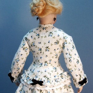 K&ouml;nnte beinhalten: Ein wei&szlig;es Kleid mit blauem Blumenmuster und schwarzen Schleifenakzenten. Das Kleid hat ein tailliertes Oberteil und einen vollen Rock mit R&uuml;schen. Die Puppe tr&auml;gt schwarze Ohrringe.