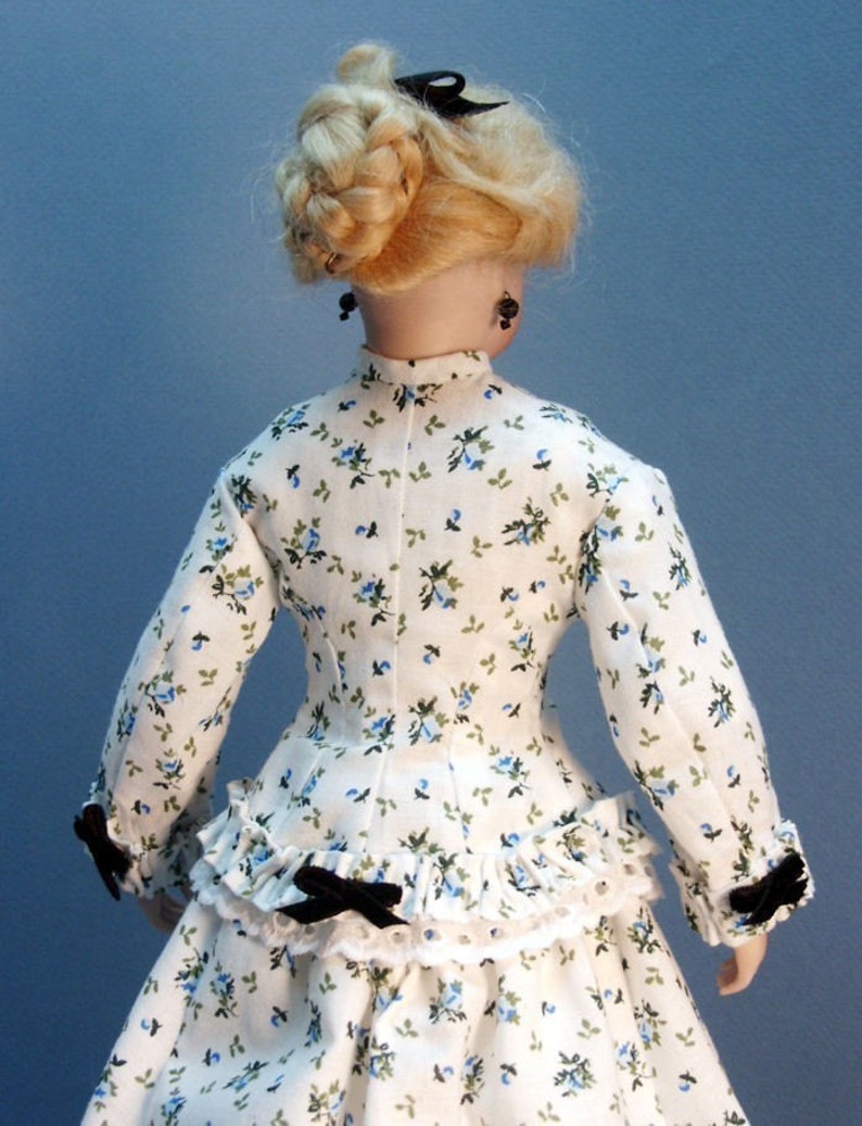 K&ouml;nnte beinhalten: Ein wei&szlig;es Kleid mit blauem Blumenmuster und schwarzen Schleifenakzenten. Das Kleid hat ein tailliertes Oberteil und einen vollen Rock mit R&uuml;schen. Die Puppe tr&auml;gt schwarze Ohrringe.